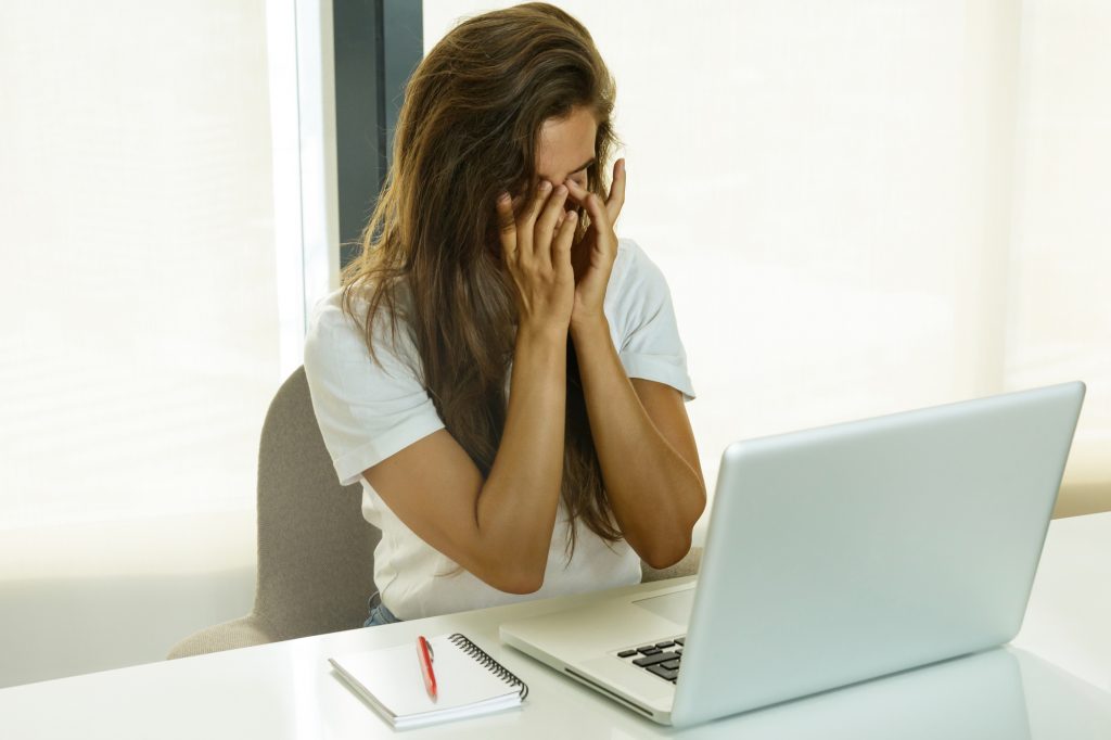 A woman is sitting in front of the laptop while touching the corner of her eyes with both hands