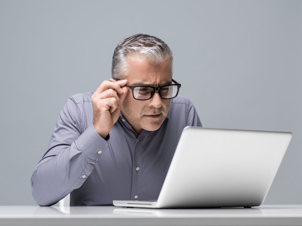 An old man holding his eyeglasses to look at the laptop in front of him