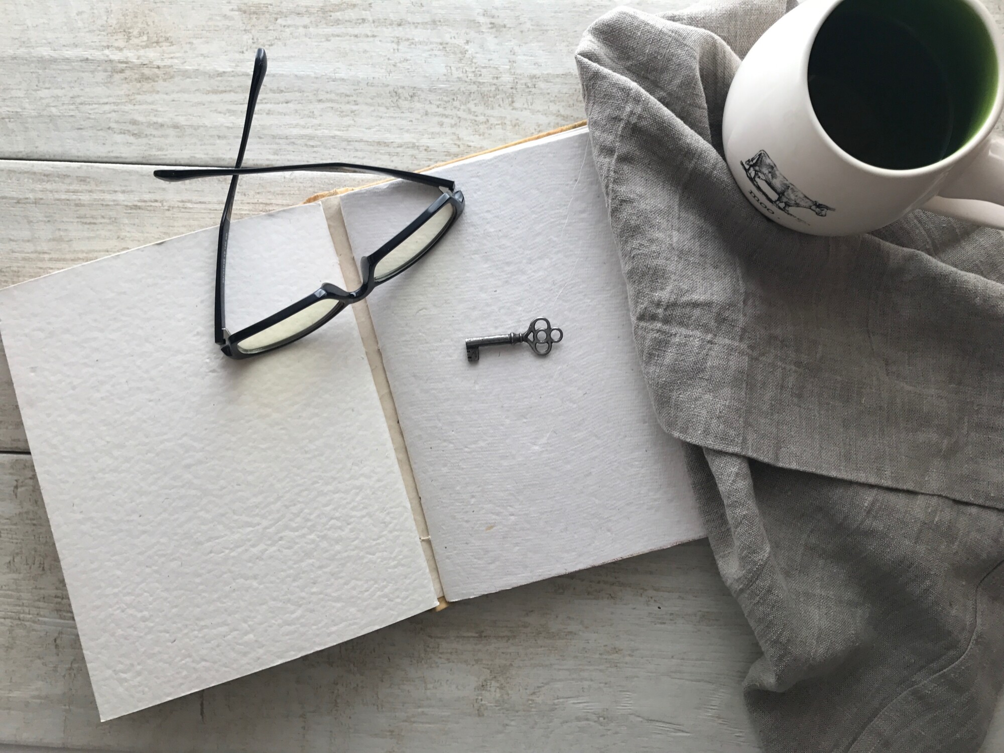 Eyeglasses on top of a blank page of a book with a key and cup of tea beside it